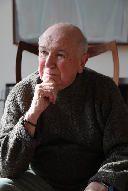 Photo of Terrence McNally, an older, balding man wearing a gray or green sweater, sitting in a chair with hist hand below his chin