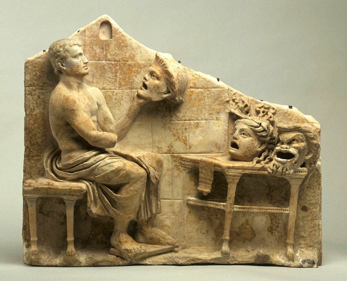 Photo of a sculpture of Thespis sitting in a chair, holding a head with two more heads on a table in front of him